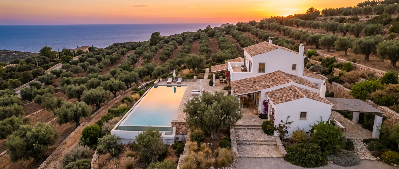 Aerial view of a white hillside villa with a pool and olive trees at sunset